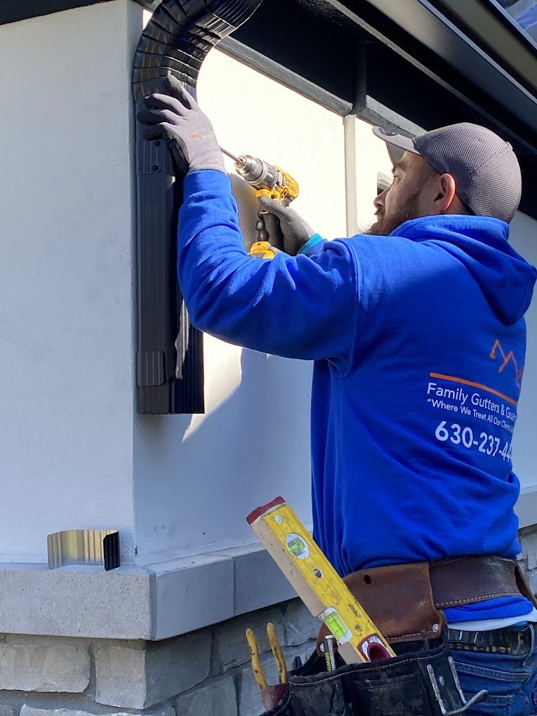 Family Gutters and Guards installer in a branded work hoodie fastening a downspout elbow to a stucco wall