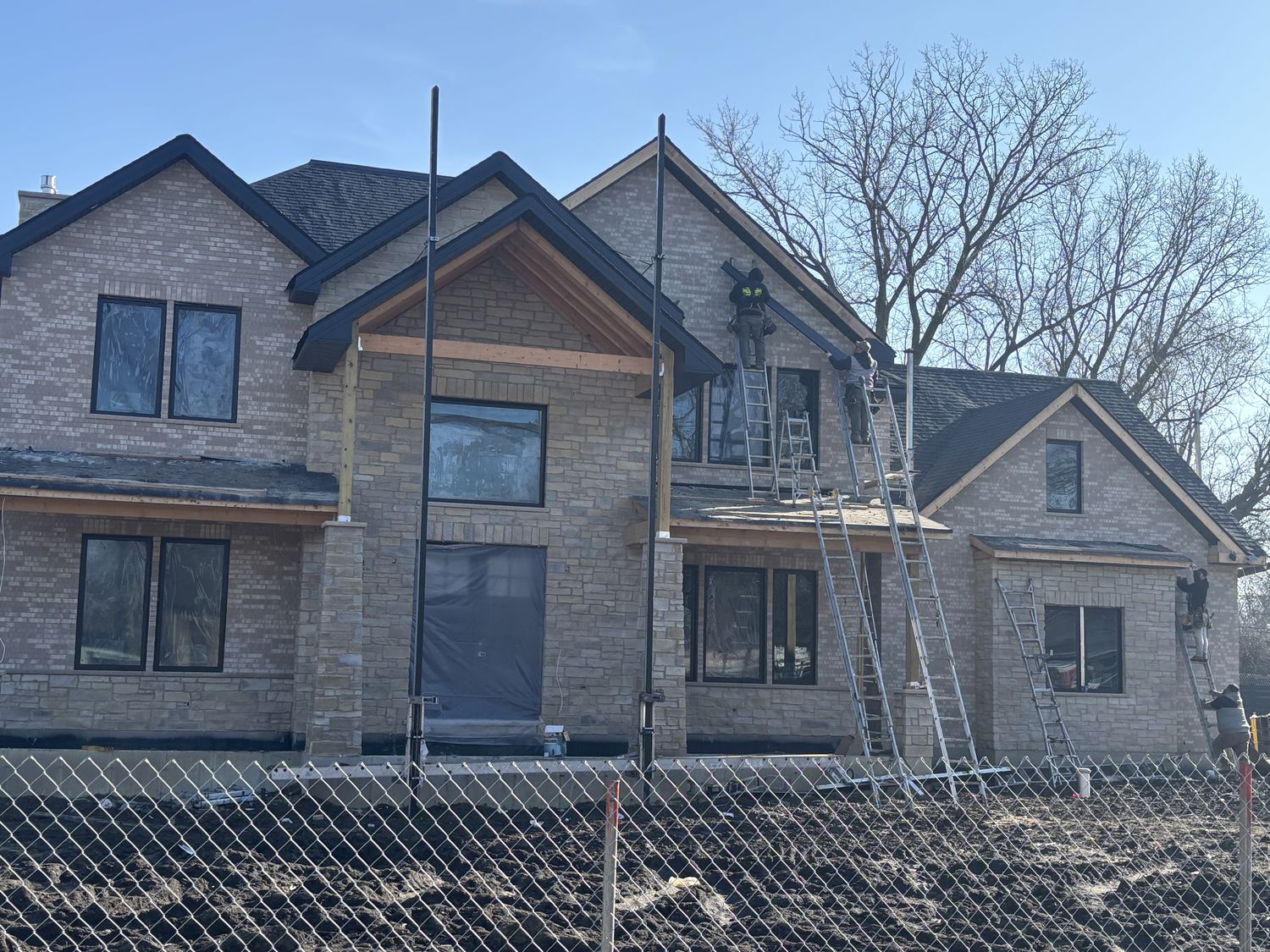 Crew on ladders installing gutters on a large brick home still under construction, dirt lot and site fence in front